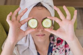 women wearing towel head wrap and robe holds up cucumber slices in front of eyes
