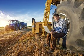 Farmer resting on a tractor wheel while working on a laptop