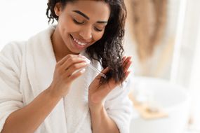 African American Woman Applying Serum On Damaged Dry Hair In Modern Bathroom At Home.
